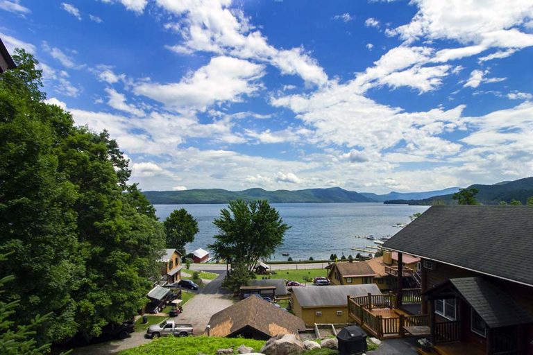 Lakeside view with blue sky, scattered clouds, calm water, tree-covered hills, houses, and a dock along a shoreline.