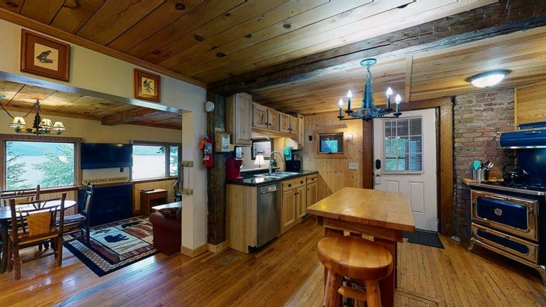 Rustic log cabin kitchen with wooden cabinets, island, chandelier, brick wall, and adjacent dining/living area.