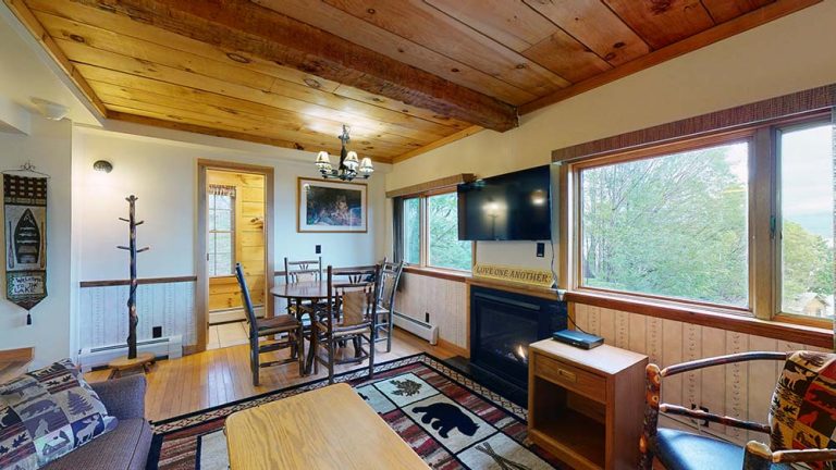 Cozy rustic living room with wooden ceiling beams, large windows, fireplace, and dining area.