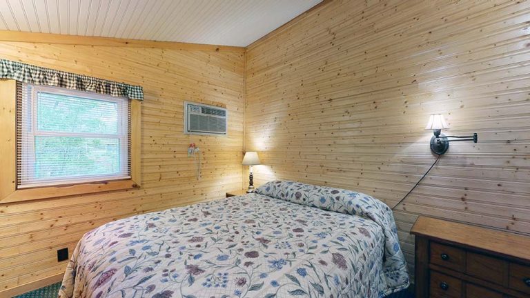Wood-paneled bedroom with window, bed, floral quilt, nightstand, and wall lamp.