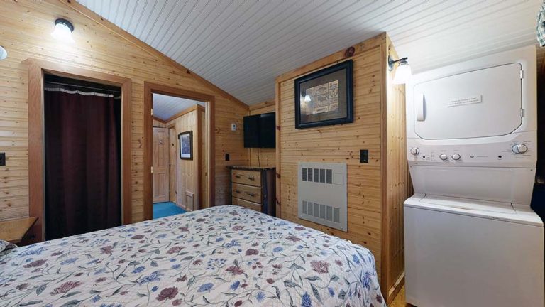 Cozy pine-paneled bedroom with floral bedspread, washer/dryer, wall TV, dresser, and doorway.