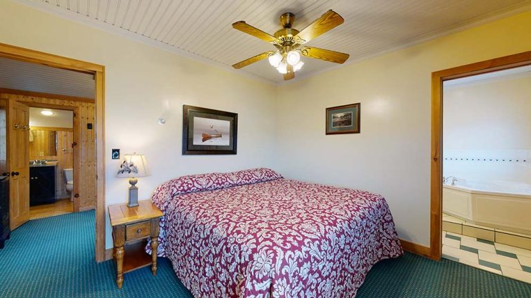 Cozy bedroom with a large bed, quilt, nightstand, lamp, and doorway to bathroom with tub.