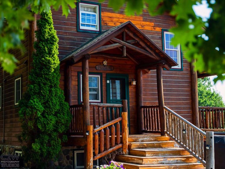 Rustic wooden two-story cabin with a covered entry porch, logs railing, and green door.