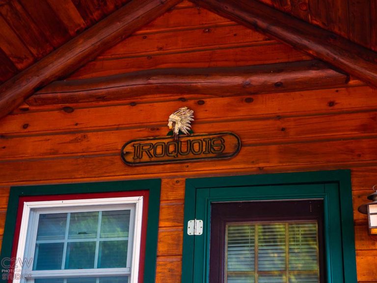Wood cabin wall with green door, white-framed window, and carved “IROQUOIS” sign above.
