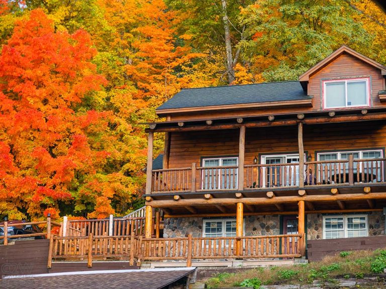 Two-story wooden cabin with stone foundation on hillside, surrounded by vibrant autumn trees.