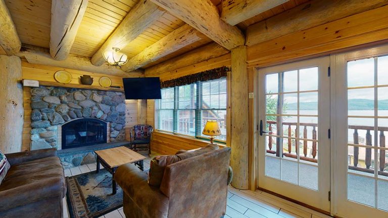 Cozy log cabin living room with stone fireplace, leather seating, and bay doors to lake view.