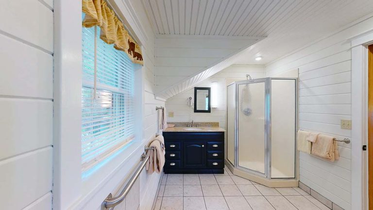 Bright white-paneled bathroom with corner shower, navy vanity, window, and towels.
