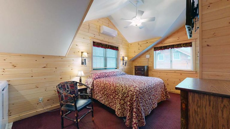 Attic bedroom with wood paneling, sloped ceiling, large bed, red patterned cover, and two windows.