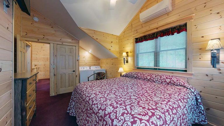 Wood-paneled bedroom with a large bed and red floral quilt, window, lamps, and dresser.