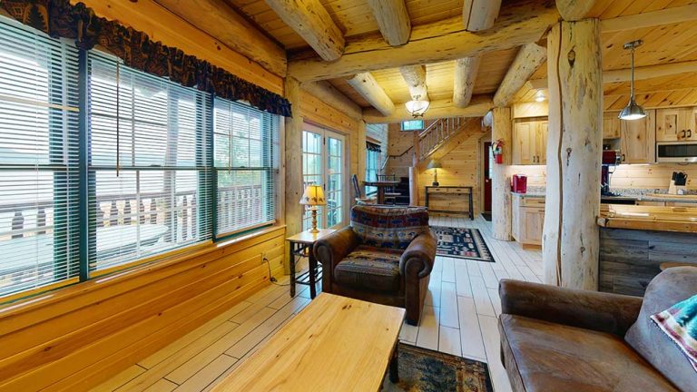 Cozy log cabin living area with wooden walls, beams, large windows, sofa, armchairs, and open kitchen.