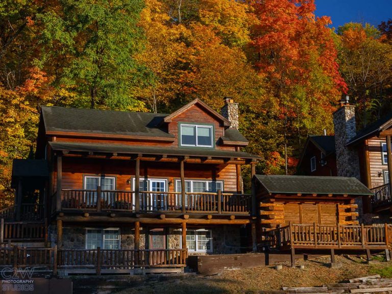 Wooden cabin with wraparound porch among autumn trees with colorful foliage.