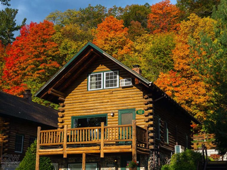Log cabin with green trim amid autumn trees, featuring a balcony and sloped roof.