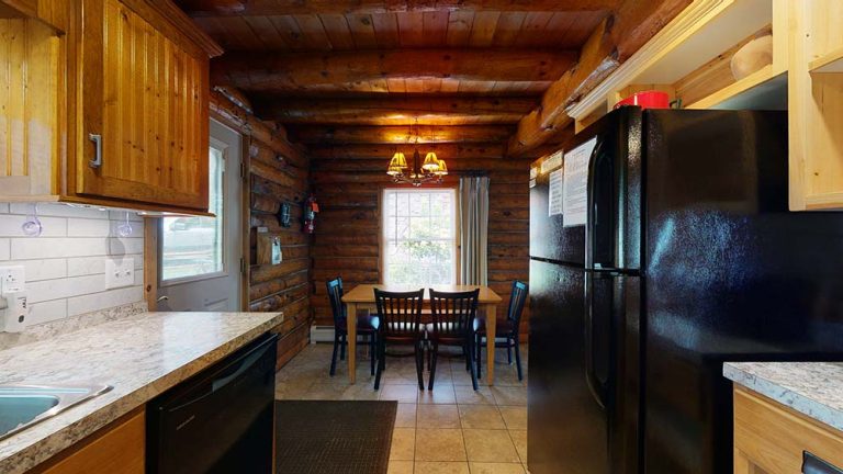 Rustic log cabin kitchen with wooden walls, dining table, six chairs, and black refrigerator.
