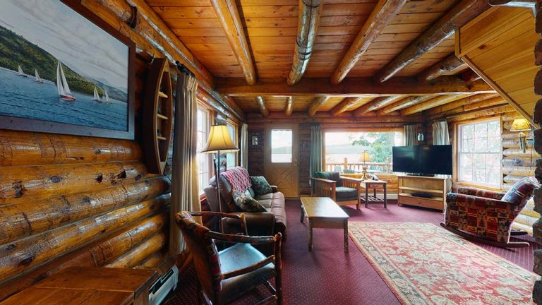 Cozy log cabin living room with wooden beams, furniture, rug, and large windows.