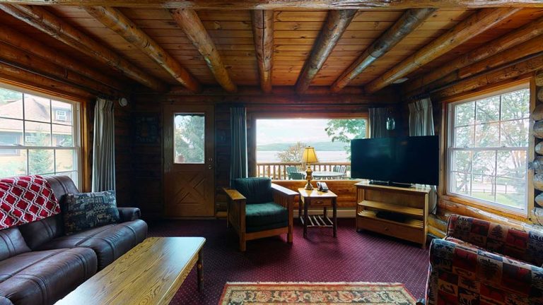 Cozy log cabin living room with leather sofa, wood furniture, and lake view through large windows.