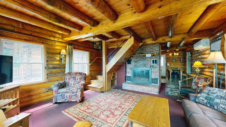 Cozy rustic living room in a log cabin with a brick fireplace, wooden beams, and stairs.