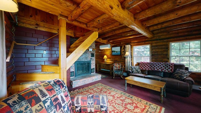 Cozy log cabin living room with exposed beams, brick fireplace, staircase, and leather sofa.