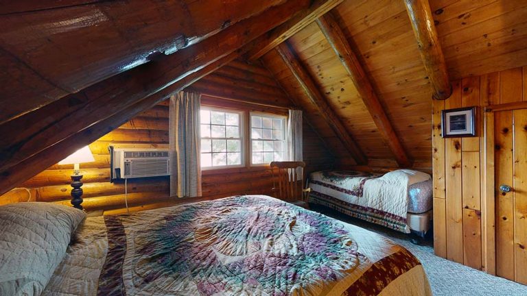 Attic bedroom with wood walls, two beds, quilted covers, window, lamp, and AC unit.
