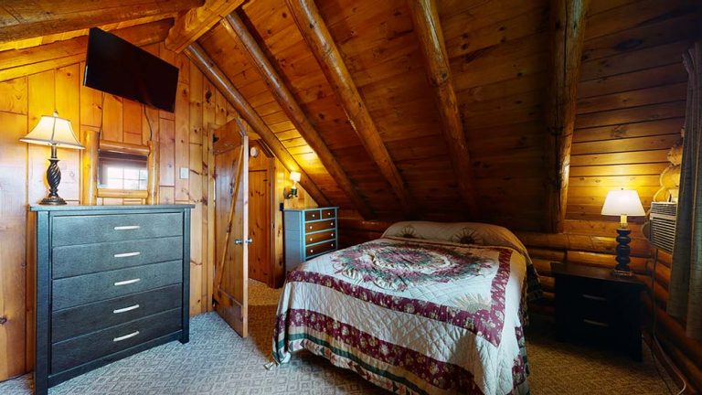 Cozy wooden attic bedroom with quilted bed, dresser, lamps, TV, and slanted beams.