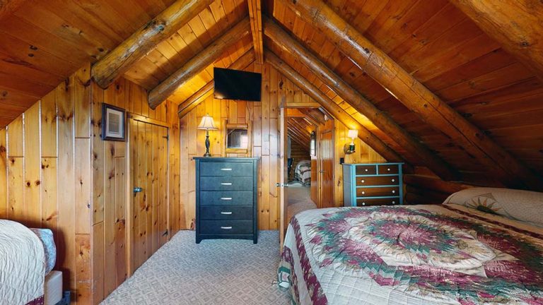 Attic bedroom with wood paneling, exposed beams, two beds, dressers, and lamps.