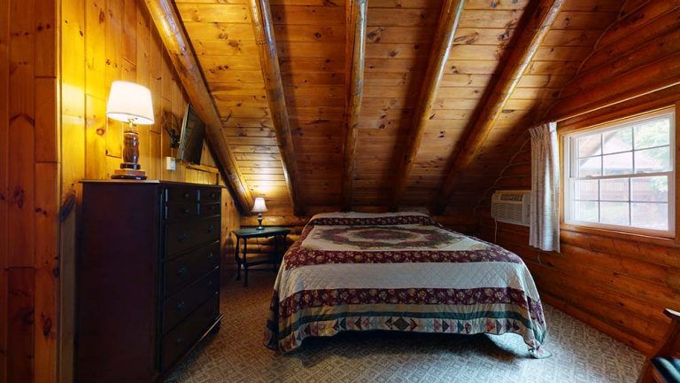 Cozy attic bedroom with exposed wooden beams, quilted bed, dresser, and window light.