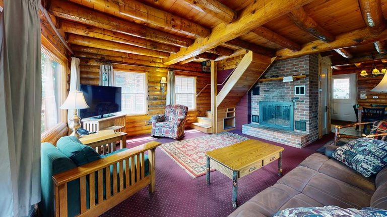 Cozy log cabin living room with wooden beams, brick fireplace, multiple seating, and warm light.