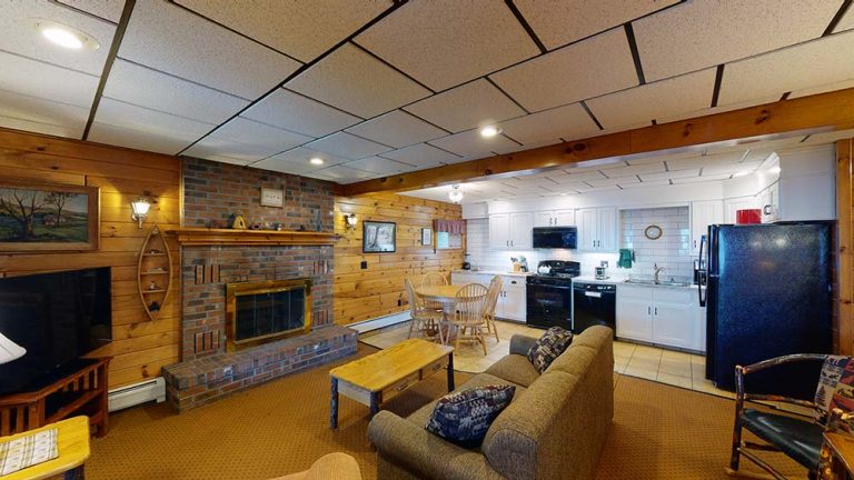Spacious rustic living area with brick fireplace, wood paneling, and an adjoining white kitchen.