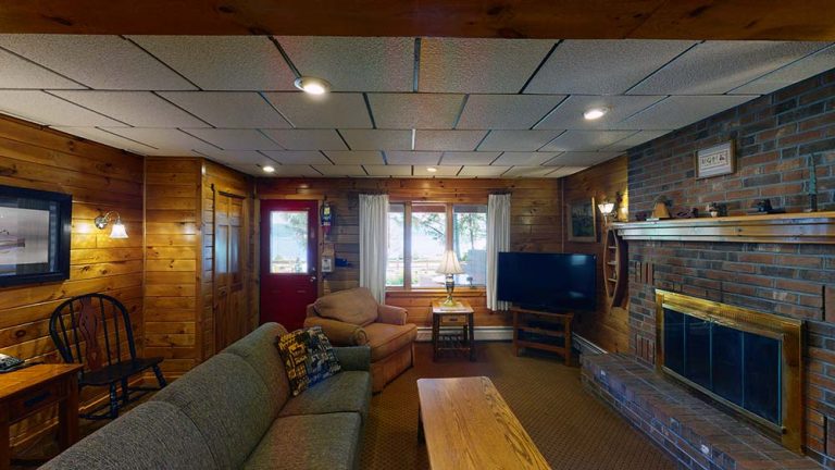 Cozy wood-paneled living room with brick fireplace, gray sofa, windows, and red front door.