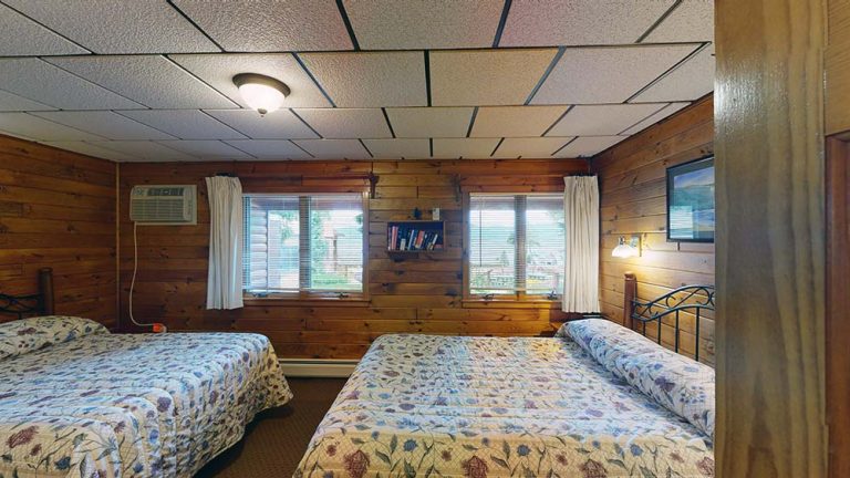 Wood-paneled motel room with two double beds, floral quilts, windows, and wall shelf.