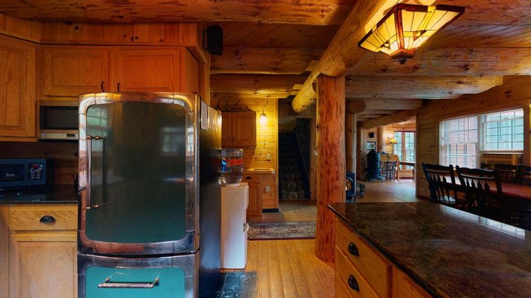 Rustic log cabin kitchen with wooden cabinets, stainless fridge, water cooler, and granite island.