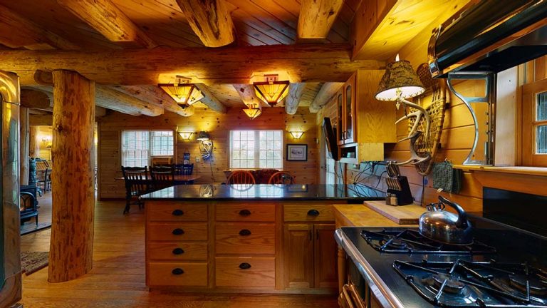Rustic log cabin kitchen with wooden beams, island, gas stove, and dining area.
