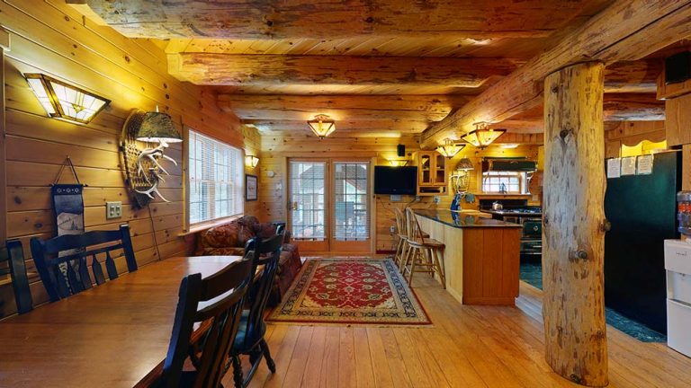 Rustic log cabin interior with dining area, sofa, kitchen island, exposed beams, and warm lighting.