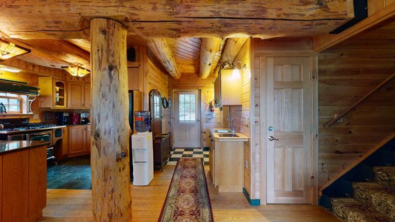 Rustic log cabin interior with wooden walls, central beam, kitchen along left, staircase on right.