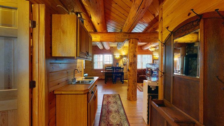 Rustic log cabin interior with small kitchen left, central wooden beam, dining area in back.
