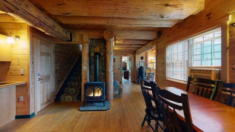 Rustic cabin interior with wooden beams, dining table, staircase, wood stove, stone chimney, and large windows.
