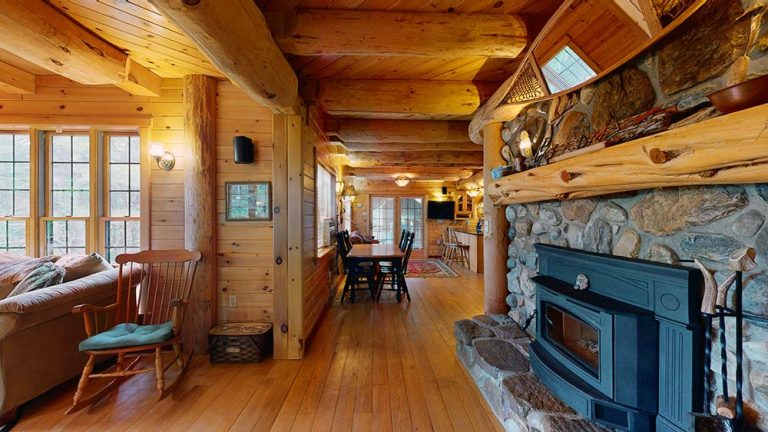 Rustic log cabin interior with stone fireplace, wooden beams, and hardwood floors; dining area center, living area left, hallway kitchen in background, large windows.