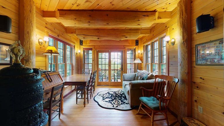 Cozy log cabin dining area with wooden beams, large windows, sofa, and rocking chair.