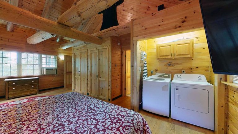 Rustic log bedroom with exposed beams, bed, wardrobes, and a nearby laundry area.