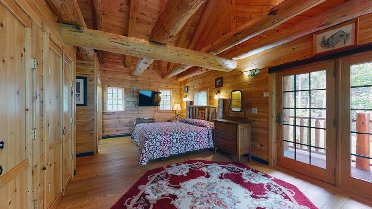 Rustic log bedroom with large bed, dresser, TV, rug, and French doors to balcony.