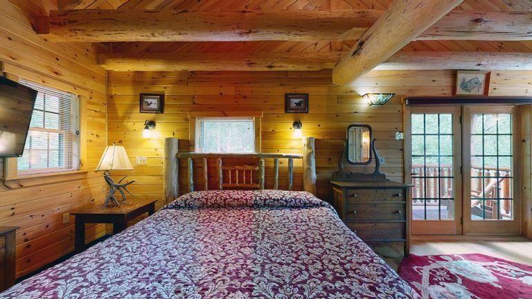 Cozy log cabin bedroom with wooden walls, large bed, burgundy patterned quilt, and glass doors to a balcony.