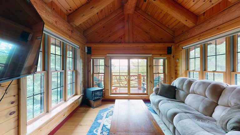 Cozy cabin living room with wooden walls, large windows, gray sectional, and door to porch.