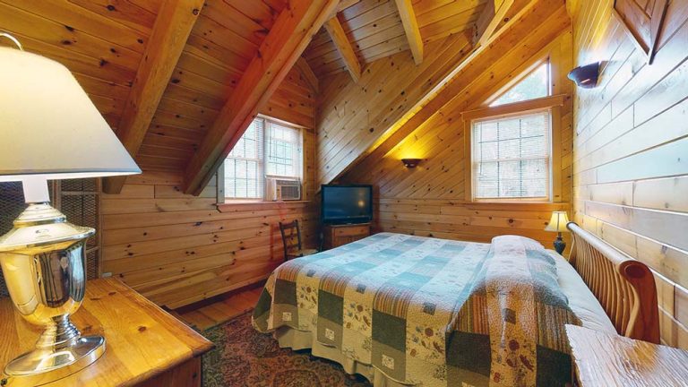 Cozy attic bedroom with wooden paneling, queen bed, quilt, dresser, TV, windows, and rug.