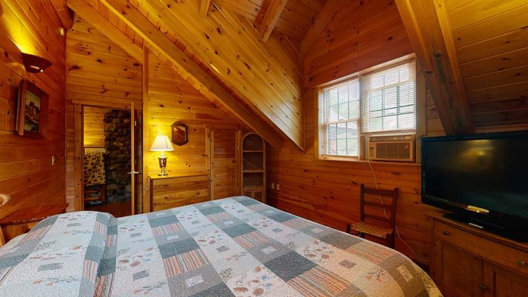 Cozy wood-paneled bedroom with patchwork quilt, dresser, window AC, and TV.
