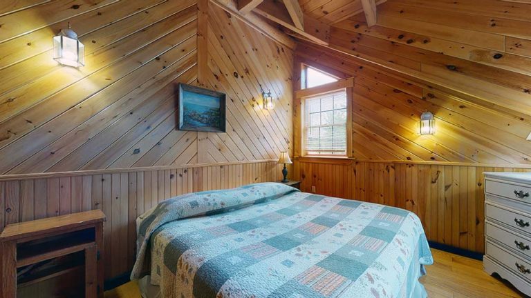Cozy sloped-ceiling bedroom with patchwork quilt, wood panel walls, window, dresser, and lamps.