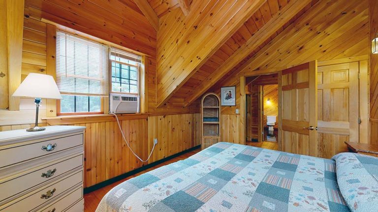 Wood-paneled cabin bedroom with two beds, dresser, lamp, window AC, and doorway to another room.