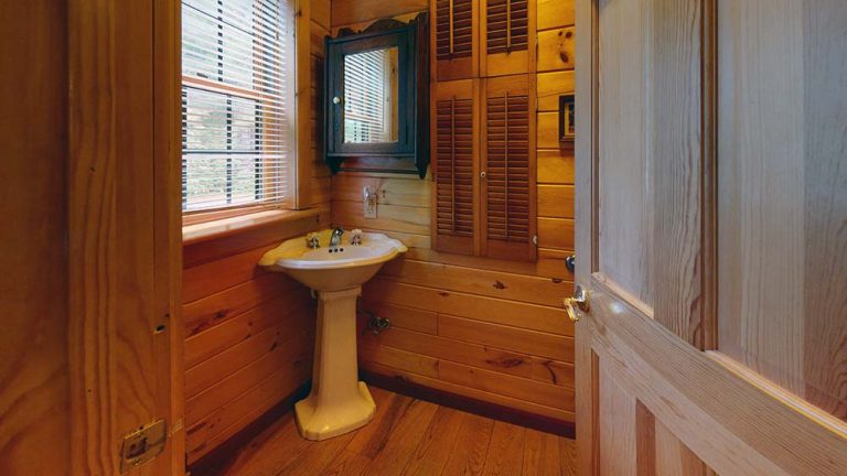 A small rustic bathroom with wooden panel walls, pedestal sink, window with blinds, and mirrored cabinet.