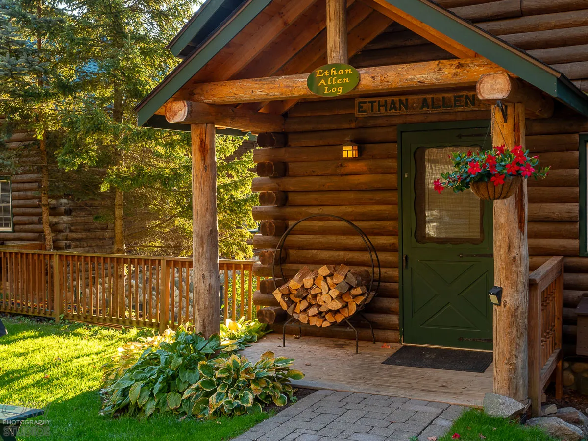 Rustic log cabin with green door, wood pile, hanging flowers, and log porch sign Ethan Allen.