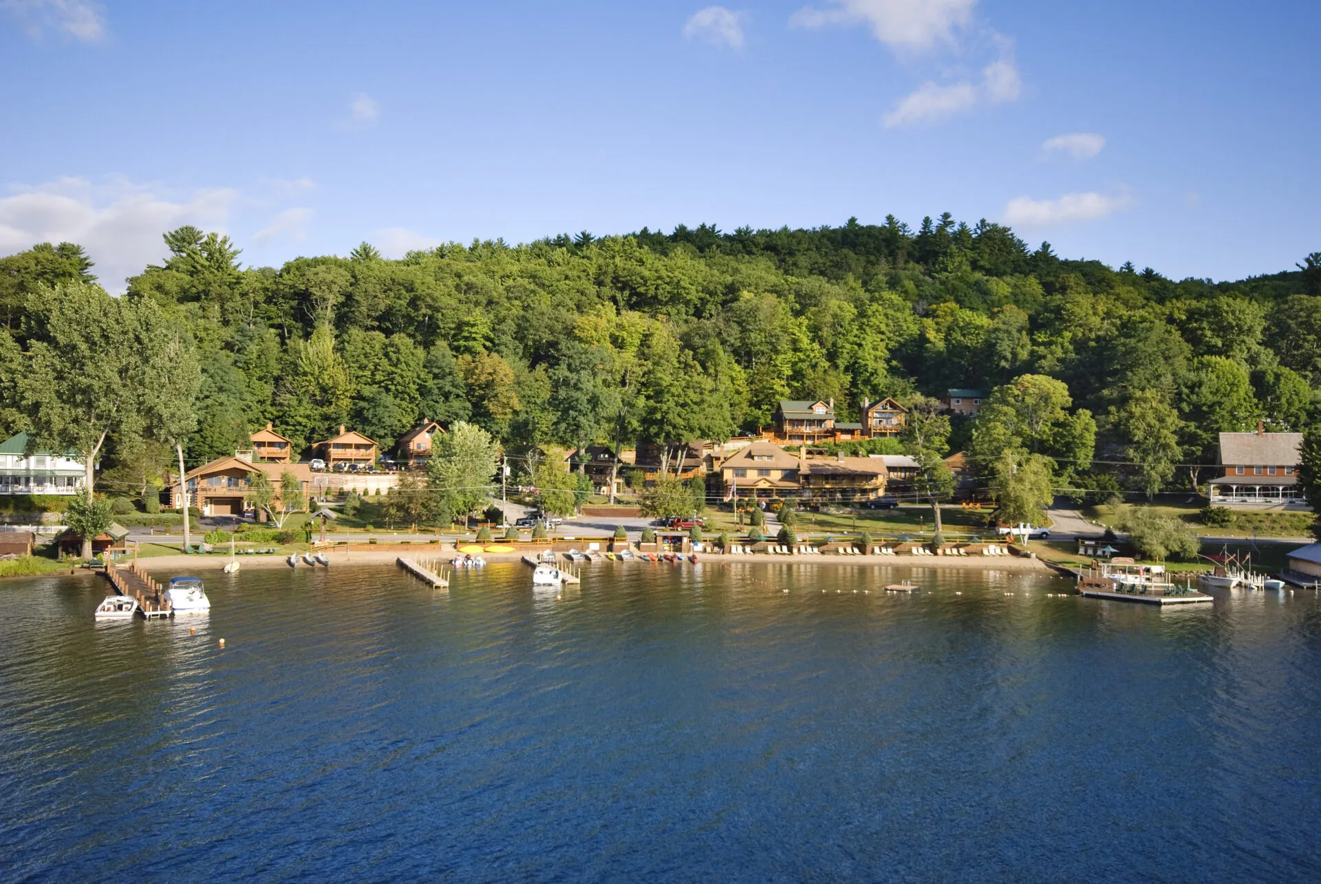 Lakeside village with wooden houses at forest edge, boats along shore, blue sky.